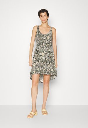 Vestido floral com uma base preta, apresentando pequenas flores rosas e amarelas, design sem mangas e uma bainha assimétrica. Combinado com sapatos de salto bloco claros.