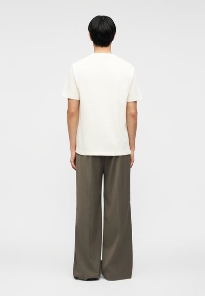 Άνδρας που στέκεται με την πλάτη προς τα εμπρός, φορώντας ένα απλό λευκό κοντομάνικο πουκάμισο και καφέ παντελόνια με φαρδιά πόδια, μπροστά σε ένα απλό φόντο.