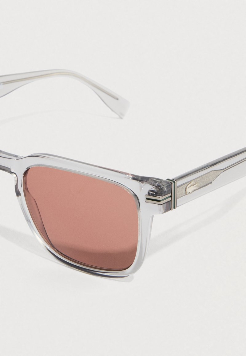 Solbriller med gennemsigtigt stel og rosatonede linser. Metalliske detaljer på stængerne med et accent ved hængslet. Rektangulært design.