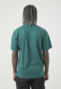 Dark green short-sleeve t-shirt with a relaxed fit, smooth texture, round neck, and straight hem. Worn with black pants.
