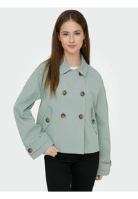 Jeune femme aux longs cheveux bruns portant une veste à double boutonnage vert clair avec de gros boutons, un pantalon noir et des boucles d'oreilles anneaux.