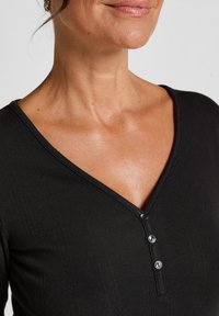 Zwarte v-hals top van textuur ribstof, met drie knopen bij de halslijn. Soepele textuur en getailleerd ontwerp.