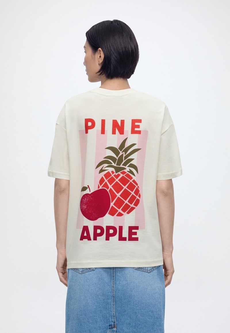 Mujer con cabello corto negro que lleva una camiseta crema con gráficos y texto en rojo de piña y manzana, combinada con una falda de mezclilla azul.