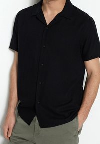 Černá košile s krátkým rukávem na knoflíky s límečkem, vyrobená z texturované látky a s volnějším střihem, doplněná olivově zelenými kalhotami.