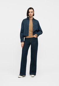 Γυναίκα που στέκεται φορώντας σκούρο μπλε πουκάμισο από τζιν, καφέ μακρυμάνικο τοπ, τζιν παντελόνι με φαρδιά μπατζάκια σε σκούρο μπλε χρώμα και μαύρα αθλητικά παπούτσια με λευκή σόλα.