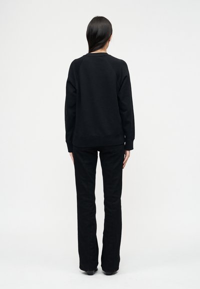 Άτομο με μακριά σκούρα μαλλιά, ντυμένο με μαύρο πουλόβερ και μαύρο παντελόνι, να στέκεται με την πλάτη προς τα εμπρός σε απλό λευκό φόντο.