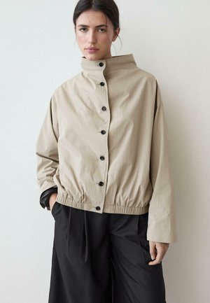 Jeune femme portant une veste beige à col montant boutonnée et un pantalon noir ample, debout avec une main dans la poche devant un mur uni.