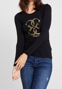 Schwarzes Langarm-T-Shirt mit einem strukturierten goldenen Logo-Druck. Weicher Stoff und figurbetontes Design. Kombiniert mit blauen Jeans.