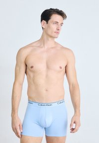 Shirtloze man met kort bruin haar in lichtblauwe Calvin Klein boxershorts, staand tegen een effen lichte achtergrond.