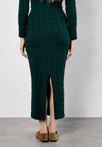 Langes grünes Strickkleid mit geripptem Oberteil, strukturiertem Muster und einem Schlitz auf der Rückseite. Kombiniert mit braunen Wildlederschuhen.