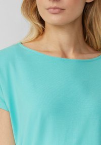 Vrouw in een lichtblauw, kort mouwtop met een brede ronde halslijn, die haar nek, kin en een deel van haar blonde haar toont.