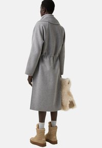 Kvinde iført en lang grå frakke og beige pelsforede støvler, der holder en beige fluffy håndtaske, vendt væk fra kameraet mod en hvid baggrund.