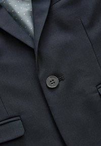 Mörk marinblå blazer med en notch-lapel, slät yta, enkel svart knappstängning och en fodring med prickar som syns inuti.