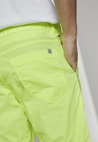 Neon žluté cargo šortky s kapsou na pravé straně a logem, vyrobené z lehké tkaniny s viditelnými švy.