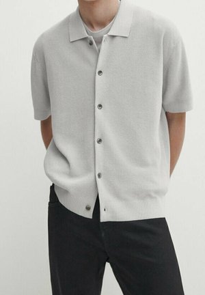 Man wearing light gray short-sleeve button-up knit shirt over a white t-shirt, paired with black pants, standing with hands behind back.