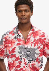 Korte mouwen shirt met een rood bloemenpatroon op een witte achtergrond, met zwarte accenten en houten knopen. Lichtgewicht stof, relaxte pasvorm.
