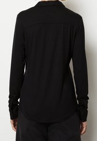 Svart långärmad skjorta med krage, med en mjuk textur, knappade ärmslut och rundad nederkant. Baksidan har en enkel design.