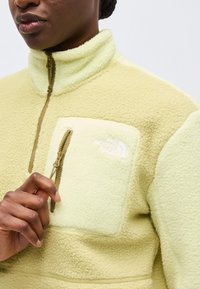 Κοντινό πλάνο ενός ατόμου που φοράει ένα ανοιχτό κίτρινο φλις μπουφάν με τσέπη στο στήθος με φερμουάρ και το λογότυπο "The North Face".