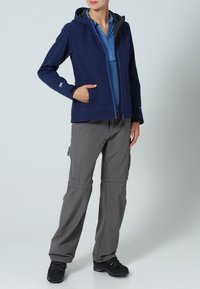 Chaqueta softshell de color marino con capucha, bolsillos frontales y cremallera morada; combinada con pantalones grises desmontables y botas de senderismo negras.