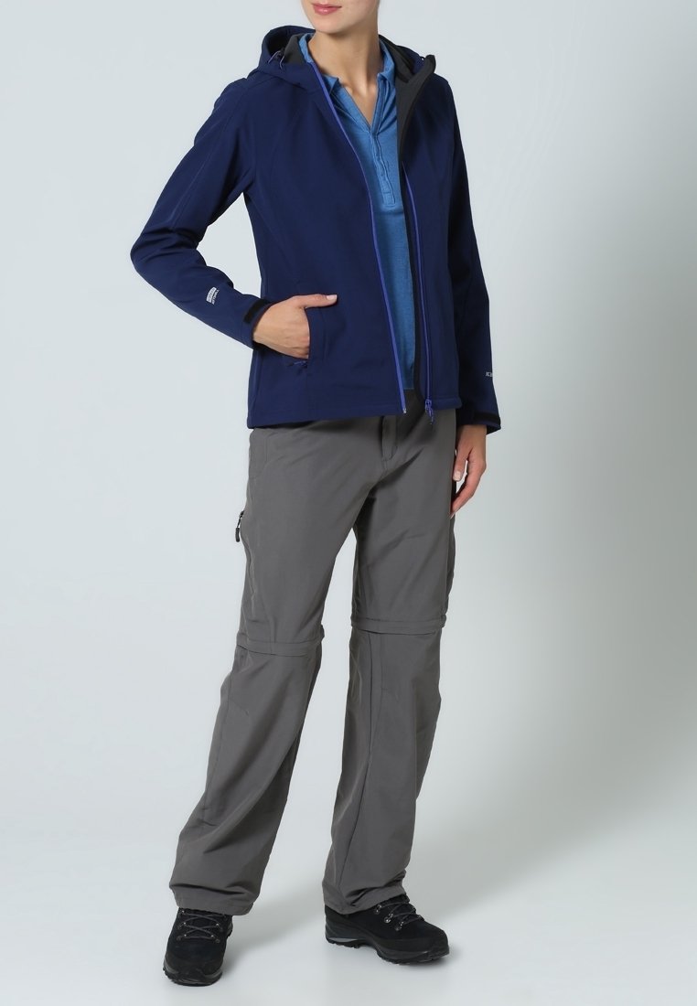 Chaqueta softshell de color marino con capucha, bolsillos frontales y cremallera morada; combinada con pantalones grises desmontables y botas de senderismo negras.