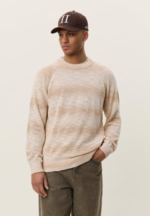 Mann trägt einen beigen Pullover und eine braune Mütze mit römischer Zahl II, steht mit einer Hand in der Tasche vor einfarbigem Hintergrund.