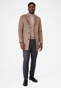 Bruine wollen blend jas met zwarte knopen, gedragen over een beige turtleneck en grijze slim-fit broek. Zwarte enkellaarsjes maken de outfit af.