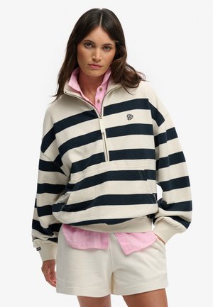 Frau trägt einen cremefarbenen und schwarzen, halb gerippten Sweatshirt über einem rosa Hemd mit Knöpfen und cremefarbenen Shorts, steht mit einer Hand in der Tasche.