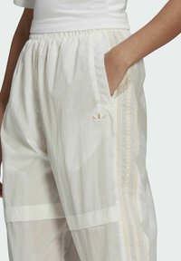 Person som bär vita Adidas träningbyxor med nitar i tre-stripesdetalj och logotyp, handen i sidofickan, tillsammans med en vit kortärmad topp.