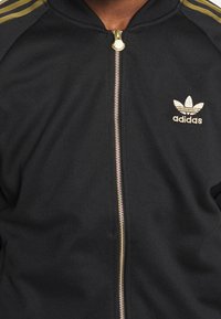 Svart zip-up jacka i texturerad tyg, med guldfärgad dragkedja och logotyp. Messingsdetaljer och tre guldstripes på axlarna.