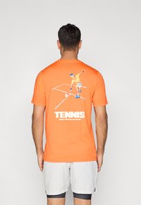 Πορτοκαλί βαμβακερό μπλουζάκι με γραφικό ενός τενίστα στην πίσω πλευρά. Διαθέτει έντονο λευκό κείμενο, και το μπλουζάκι έχει στρογγυλή λαιμόκοψη και κοντά μανίκια.