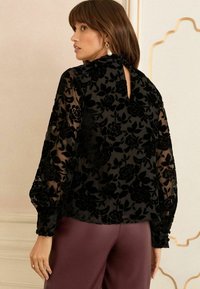 Černá blůza s květinovým vzorem a průhlednými dlouhými rukávy, vysokým límcem a detailem na zádech ve tvaru klíčové dírky, v kombinaci s bordovými kalhotami. Měkká textura.