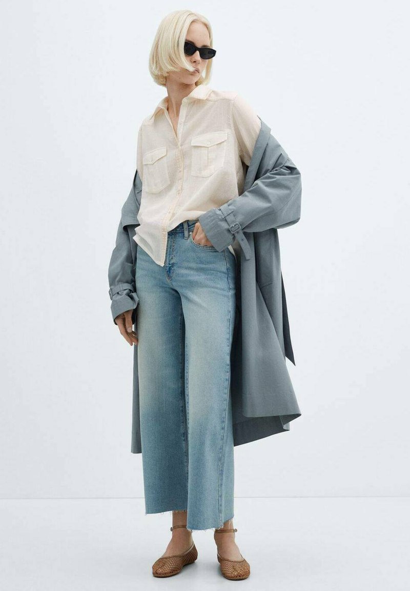 Camicia button-up beige chiaro con due tasche sul petto, abbinata a jeans blu chiaro larghi e un cappotto grigio. Scarpe intrecciate marroni completano il look.