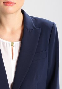 Námořnický blazer s ostrými klopami, vybavený jednou kapsou na prsou, vrstvený přes bílou halenku s výstřihem ve tvaru klíčové dírky a zlatým kováním.