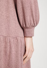 Robe en maille rose pâle à manches longues avec poignet froncé, présentant un gros plan sur la manche et une main détendue sur le côté.