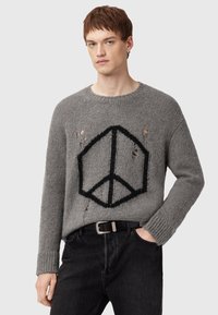 Giovane uomo che indossa un maglione grigio lavorato a maglia con un simbolo della pace geometrico nero e buchi strappati, abbinato a jeans neri e una cintura.