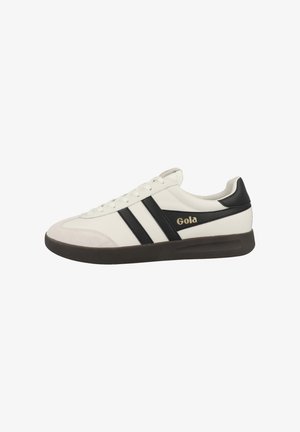 Zapatilla blanca con rayas negras y una puntera de ante. Presenta el logo de Gola en dorado en el lateral y una suela de goma oscura. Diseño atlético clásico.