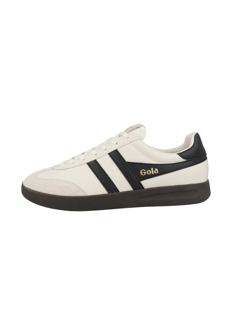 Zapatilla blanca con rayas negras y una puntera de ante. Presenta el logo de Gola en dorado en el lateral y una suela de goma oscura. Diseño atlético clásico.