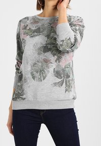 Kobieta ubrana w szary sweatshirt z subtelnym zielono-różowym tropikalnym wzorem liści, w połączeniu z ciemnoniebieskimi dżinsami i kolczykami w kształcie kół.