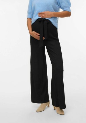 Schwarze weit geschnittene Hose aus strukturiertem Stoff mit einem gebundenen Taillenbund. Kombiniert mit einem weichen blauen kurzärmeligen Oberteil und beigen Absatzschuhen.