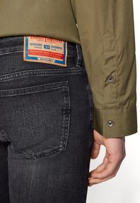 Camisa verde oliva con puños abotonados, llevada sobre unos vaqueros gris oscuro que presentan una etiqueta marrón de Diesel en el bolsillo trasero. Con líneas limpias y un diseño ajustado.