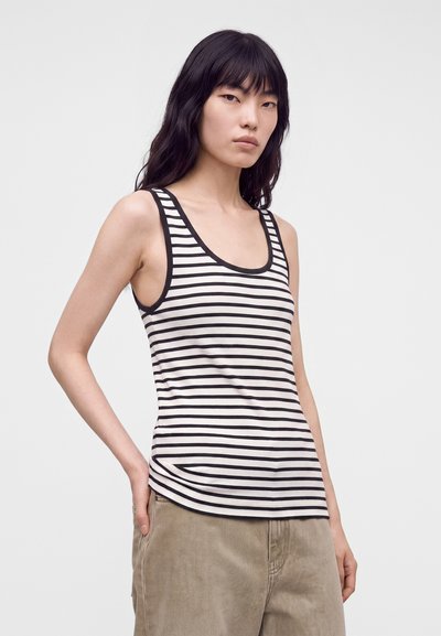 Jeune femme aux longs cheveux noirs portant un haut sans manches rayé noir et blanc et un pantalon beige, debout avec une main dans la poche.