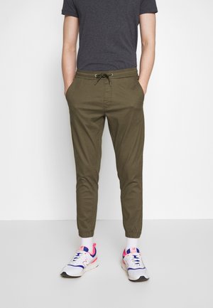 Person trägt ein dunkelgraues T-Shirt, olivgrüne Jogginghose mit Kordelzug, weiße Socken und weiße Sneakers mit blauen und rosa Akzenten.