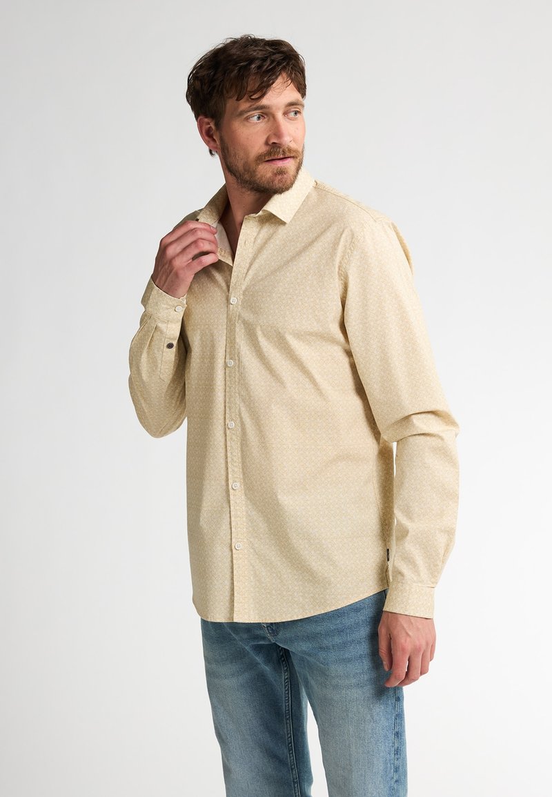 Camisa de botones beige con un patrón sutil, de manga larga y cuello clásico, combinada con jeans de color azul claro.