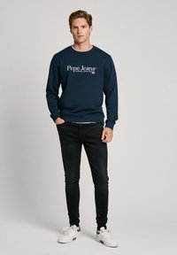 Tamno plavi dukserica s bijelim logotipom "Pepe Jeans", u kombinaciji s crnim uskim trapericama i bijelim tenisicama s plavim detaljima.