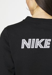 Žena v černé mikině Nike s velkým bílým vyšitým logem na zádech a viditelným stříbrným kruhovým náušnicí.