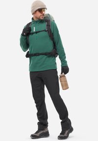 Grüner Pullover, schwarze Hose und Handschuhe. Rucksack mit grünen Akzenten, braune Wasserflasche. Graue Stiefel mit schwarzer Sohle. Reflektierende Sonnenbrille.