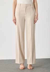 Beige wijde broek met een gladde textuur, voorzien van voorplooien, een hoge taille en subtiele stiksels. Gecombineerd met lichte sandalen.