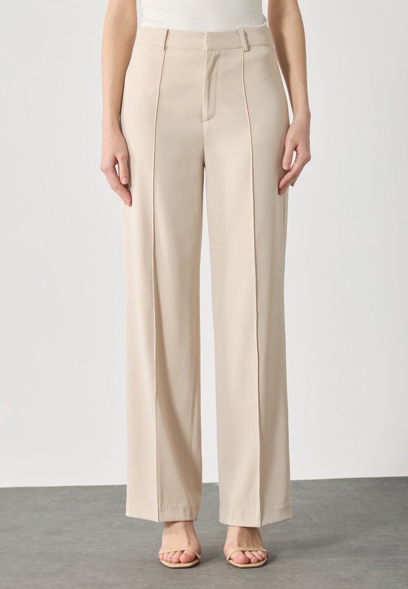 Beige wijde broek met een gladde textuur, voorzien van voorplooien, een hoge taille en subtiele stiksels. Gecombineerd met lichte sandalen.