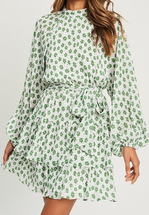 Kobieta w białej sukience z długim rękawem z zielonym kwiatowym nadrukiem, z falbanami na spódnicy i wiązaniem w talii, przedstawiona od ramion do połowy uda na neutralnym tle.