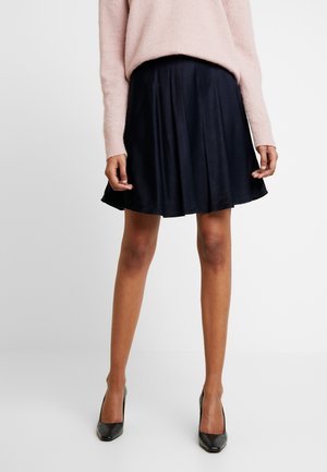 Femme portant un pull rose, une jupe évasée noire et des chaussures à talons noires, debout sur un fond blanc uni.